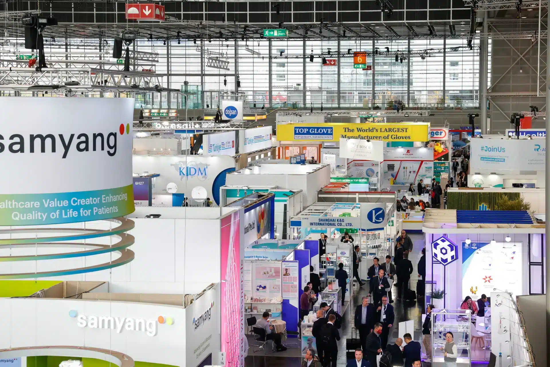 Auf der belebten Messefläche der Medica Düsseldorf präsentieren zahlreiche Stände Marken aus dem Gesundheits- und Medizinbereich. Besucher in Business-Kleidung knüpfen Kontakte und knüpfen Kontakte. Auf Bannern über den Köpfen prangen stolz Firmenlogos wie „Samyang“ und „Top Glove“ und unterstreichen die globale Reichweite der Veranstaltung.