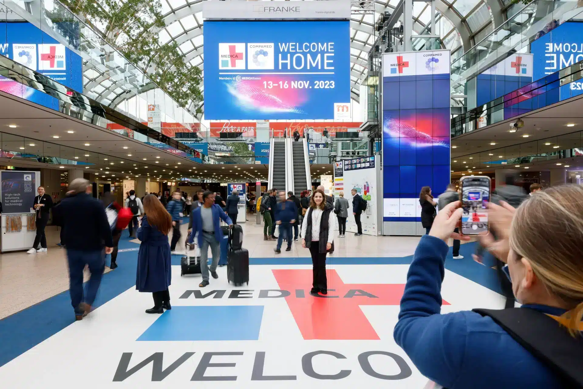 In der belebten Lobby des Düsseldorfer Kongresszentrums strömen Menschen unter einem großen Banner mit der Aufschrift „Willkommen zu Hause, 13.–16. November 2023, Medica“ hindurch. Ein Besucher fotografiert einen anderen Besucher, der auf der auffälligen Bodengrafik mit der Aufschrift „Medica Welcome“ posiert.