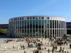 Unter strahlend blauem Himmel drängen sich Menschenmassen vor dem modernen, verglasten Kongresszentrum Messe Berlin. Dieser ikonische Veranstaltungsort mit seiner runden Struktur und den hohen weißen Säulen trägt stolz seinen Namen an der Spitze.