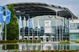 Außenansicht der Münchner Messe mit einer modernen Glas- und Stahlkonstruktion. Mehrere Flaggen säumen den Gehweg und spiegeln sich in einem Teich im Vordergrund. Ein Vogelschwarm fliegt an einem leicht bewölkten Tag über den Himmel.