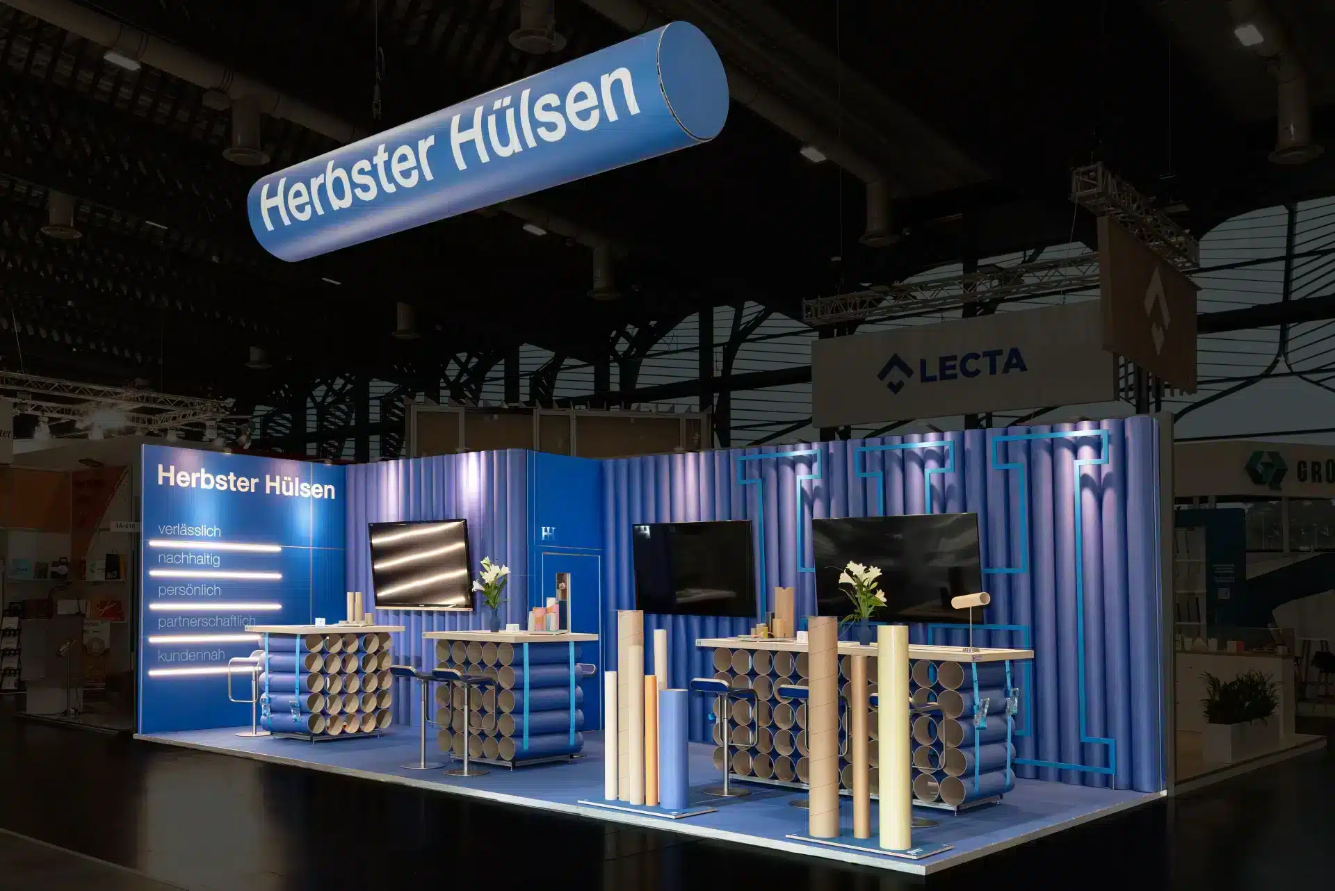 Beleuchteter Messestand von Herbster Hülsen auf der FachPack 2024 in Nürnberg mit großen Displays und dekorativen Papprohren.