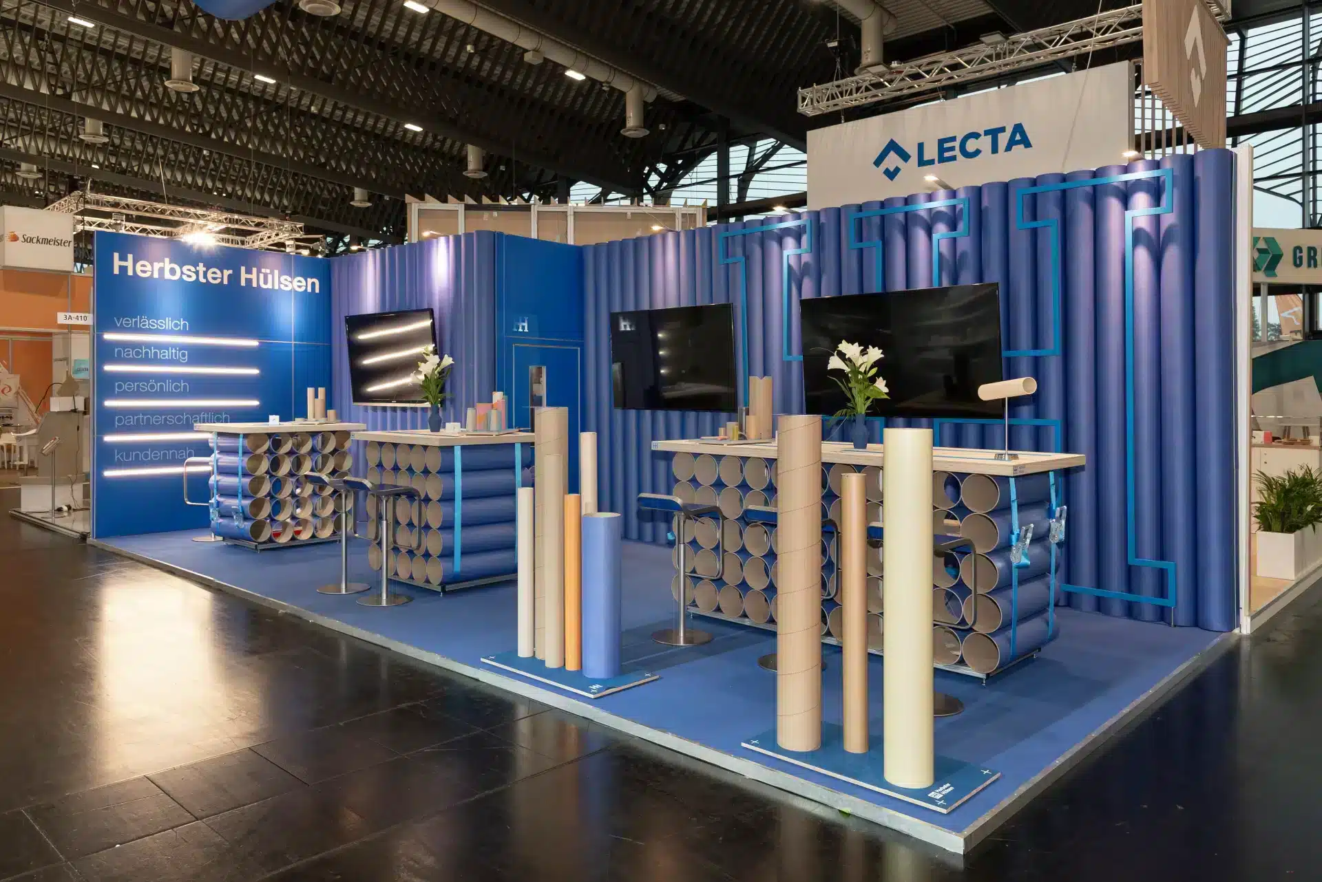 Moderner Messestand von Herbster Hülsen auf der FachPack 2024 in Nürnberg mit blauer Rückwand, beleuchtetem Slogan und Präsentationstischen aus Papprollen.