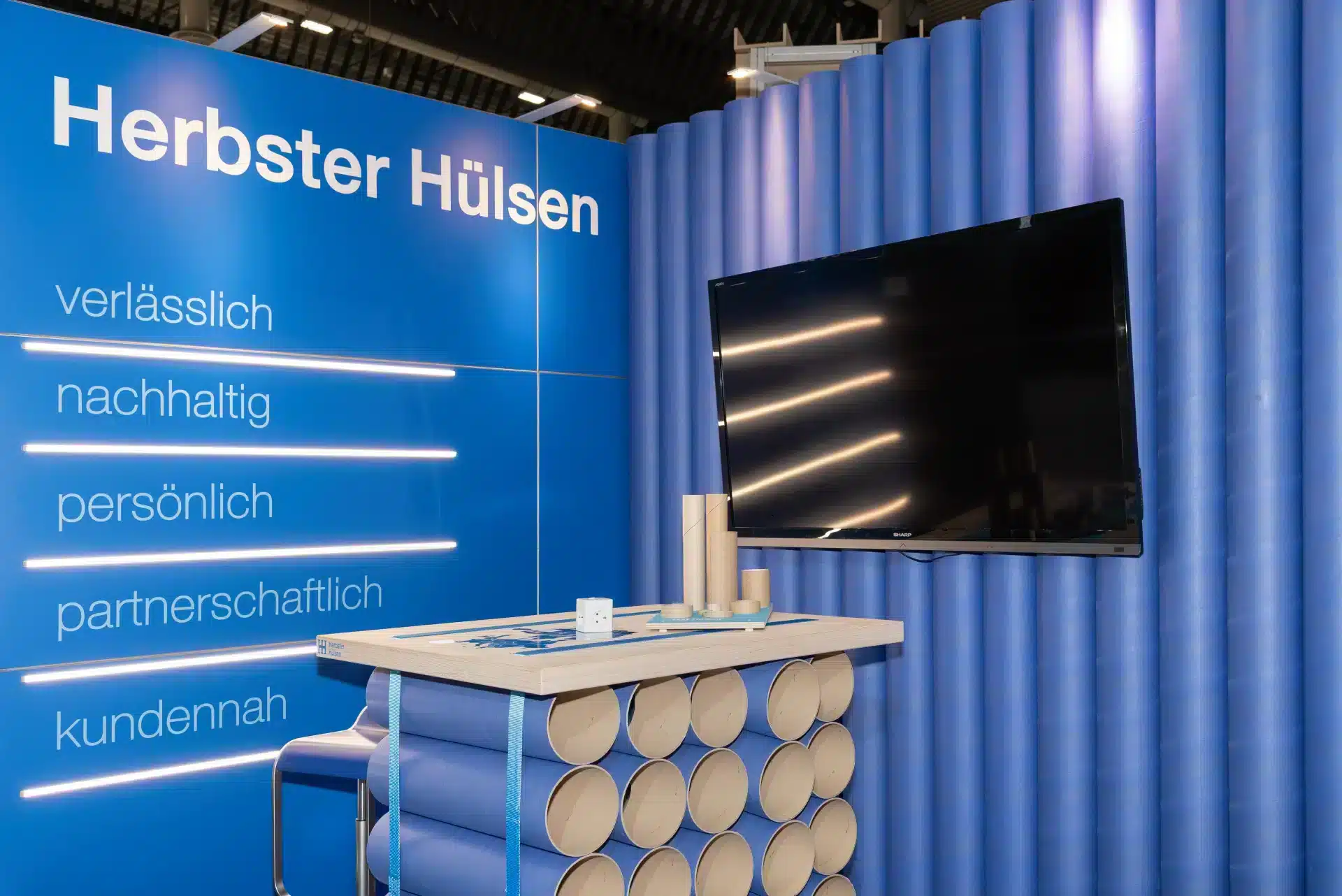 Ausstellungsstand im blauen Design mit gestapelten Pappröhren als Dekoration. An der Wand ist ein großer Bildschirm montiert. Der Text an der Wand enthält „Herbster Hülsen“ und mehrere deutsche Wörter: „verlässlich“, „nachhaltig“, „persönlich“, „partnerschaftlich“, „kundennah“.