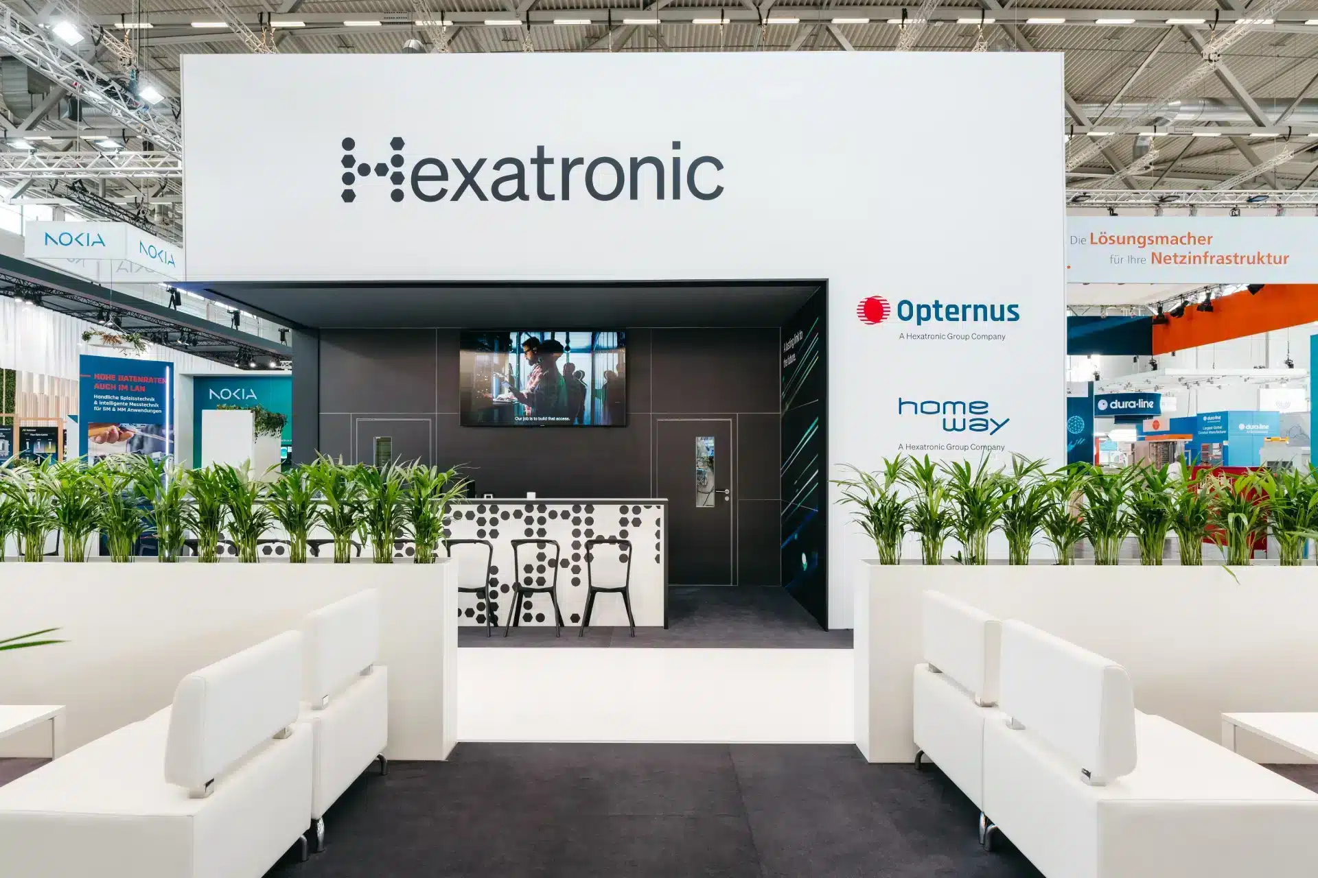 Frontalansicht des Hexatronic Messestandes mit Logo, grüner Bepflanzung und schwarzer Theke.