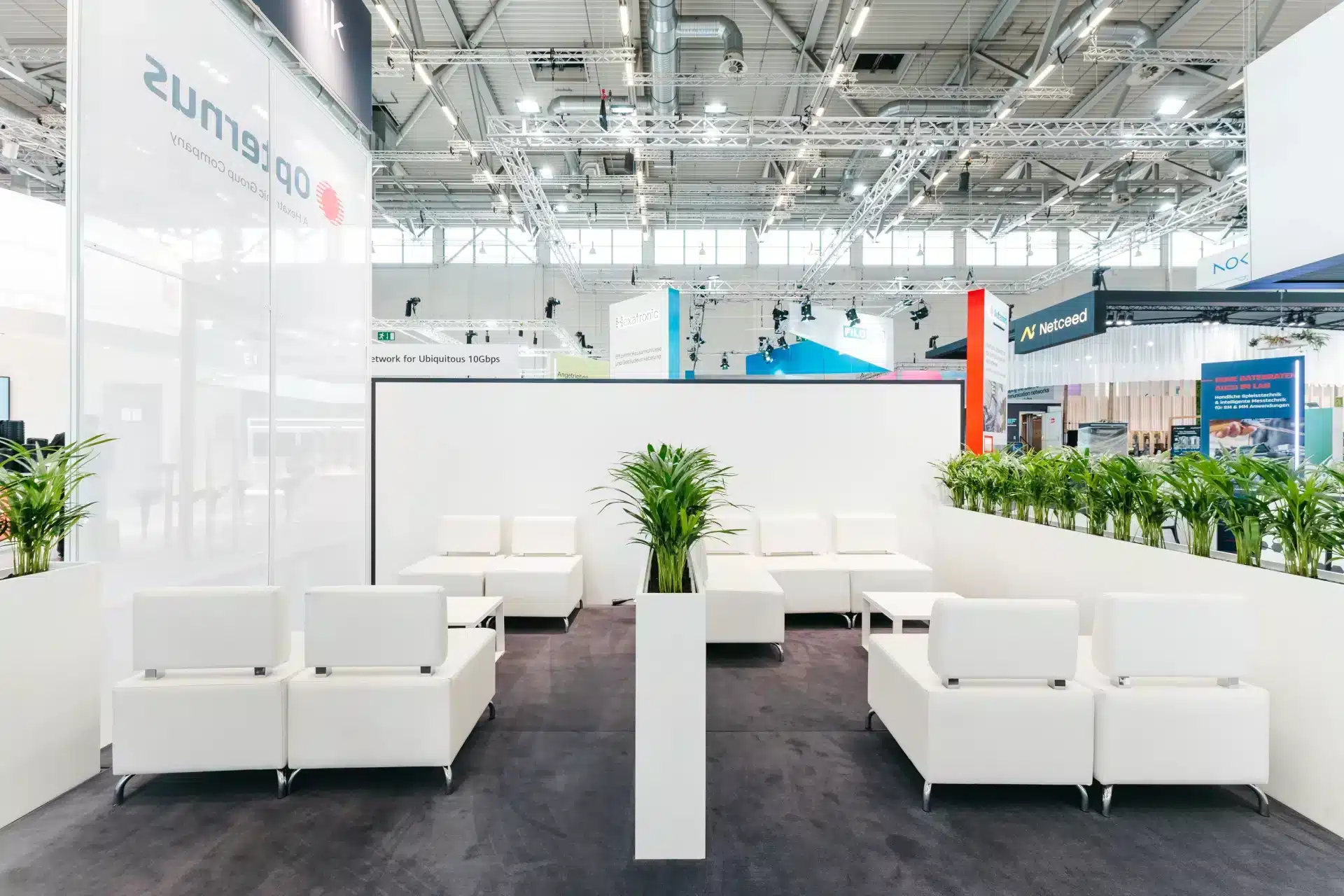 Weißer Lounge-Bereich mit modernen Sitzmöbeln und Pflanzen im Messestand von Hexatronic und Opternus.