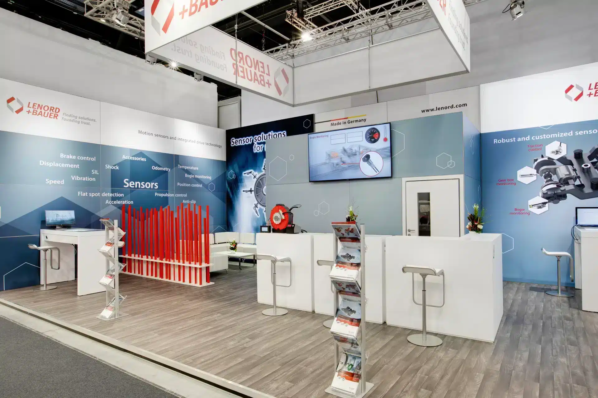 Blick auf den Lenord + Bauer Messestand mit roter Stangeninstallation, Infotheke und großem Bildschirm.