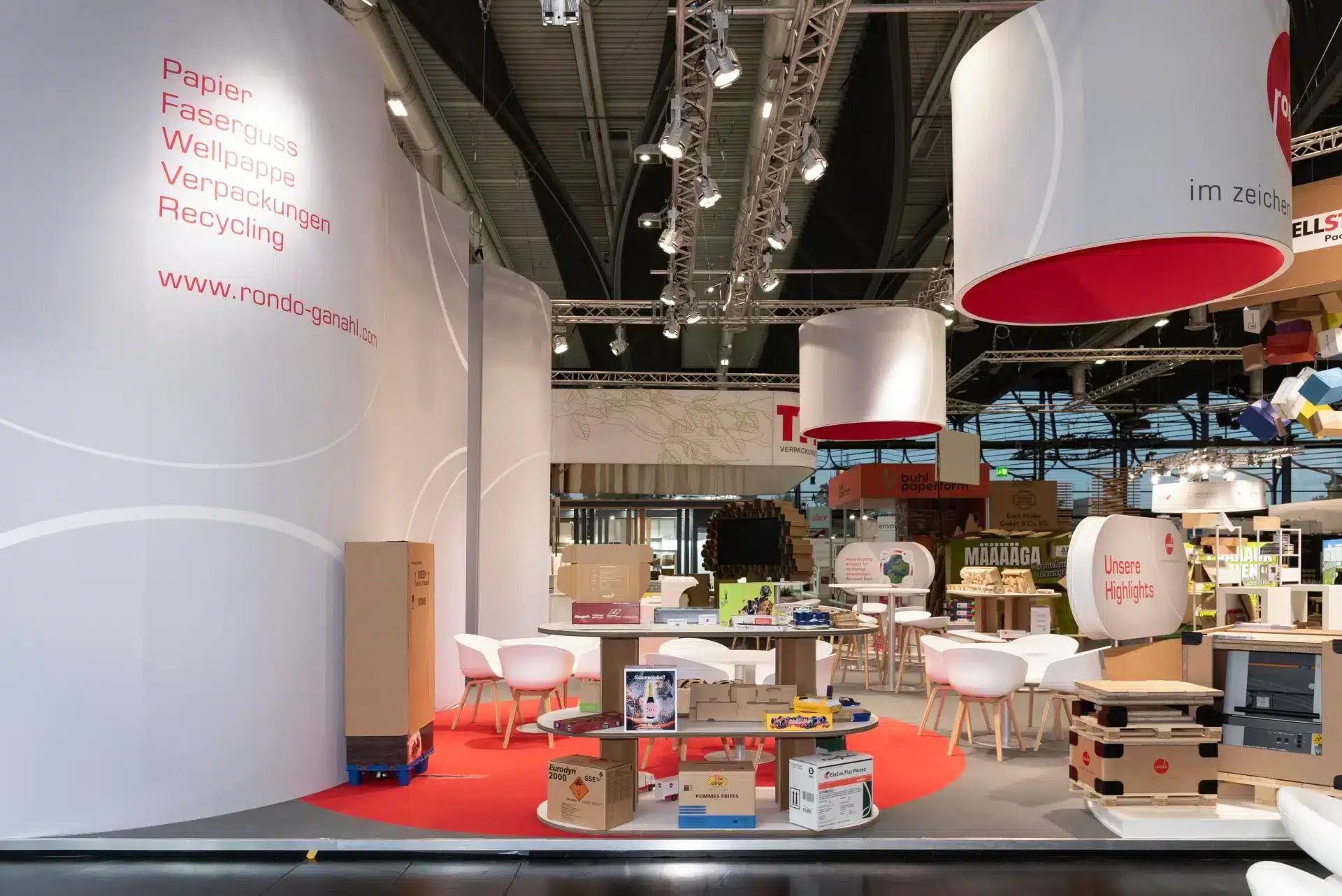 Seitenansicht des Rondo Messestandes mit roten Teppichen, weißen Sitzmöbeln und ausgestellten Verpackungen aus Papier, Wellpappe und Recycling – Messebau Nürnberg.