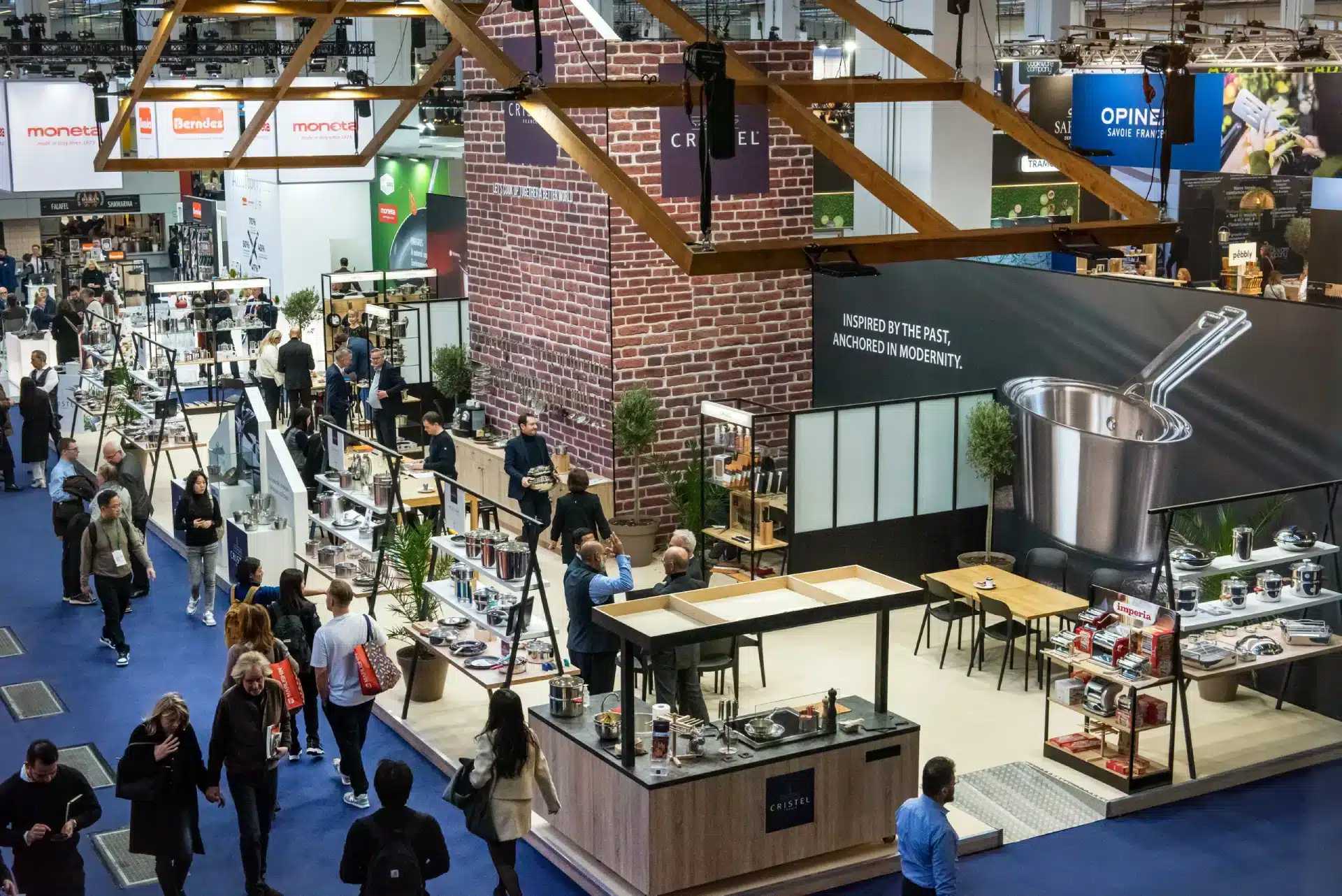 Eine geschäftige Messe, auf der Besucher bei stimmungsvoller Beleuchtung verschiedene Stände mit Küchenutensilien erkunden. Zu den Ausstellungsstücken gehören Kochgeschirr, Küchenutensilien und Dekoration. Ein zentraler Stand zeigt prominent einen Kochtopf. Die Atmosphäre ist lebhaft und professionell.