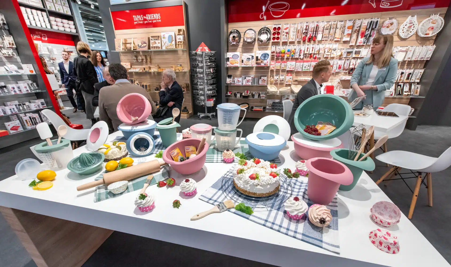 Eine bunte Auswahl an Küchenutensilien, darunter Schüsseln, Nudelhölzer und Backzubehör, sorgt für eine lebendige Atmosphäre auf dem Tisch mit Cupcakes und Kuchen. Dahinter versammeln sich die Menschen in einem geschäftigen Laden mit Regalen voller vielfältiger Küchenprodukte.