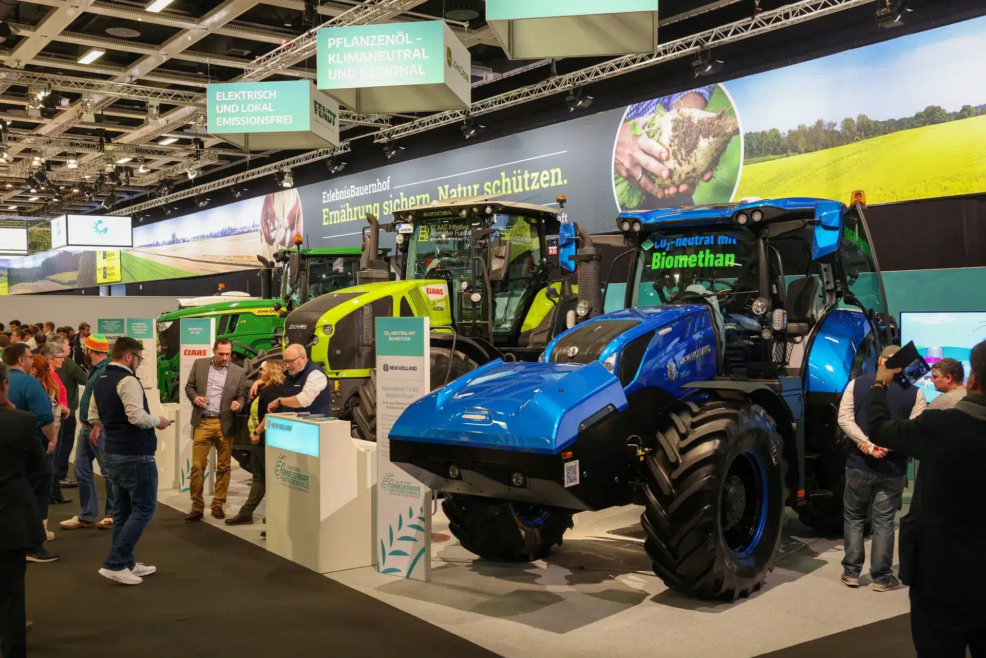 Auf der Grünen Woche versammeln sich die Besucher um große landwirtschaftliche Traktoren im Innenbereich. Um die Traktoren mit der Aufschrift „Biomethan“ herum stehen Informationsstände. Darüber heben Transparente die umweltfreundliche Landwirtschaft und den Einsatz von Pflanzenöl hervor.