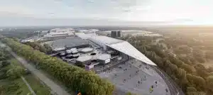 Luftaufnahme des großen, modernen Messegeländes der Messe Düsseldorf, umgeben von Bäumen. Das Gebäude zeichnet sich durch ein markantes dreieckiges Glasdach aus. Angrenzende Parkplätze sind sichtbar, und ein klarer Himmel erstreckt sich über die Stadtlandschaft und das Grün.