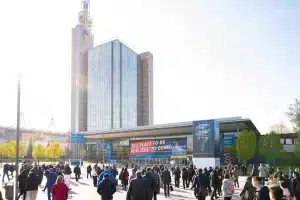 Eine Menschenmenge marschiert auf das Messegelände Hannover zu, umgeben von hohen Gebäuden aus Glas und Backstein unter klarem Himmel. Auf Bannern sind Grafiken mit Tech-Motiven und der Slogan „The Place to Be for Tech to Come“ zu sehen.