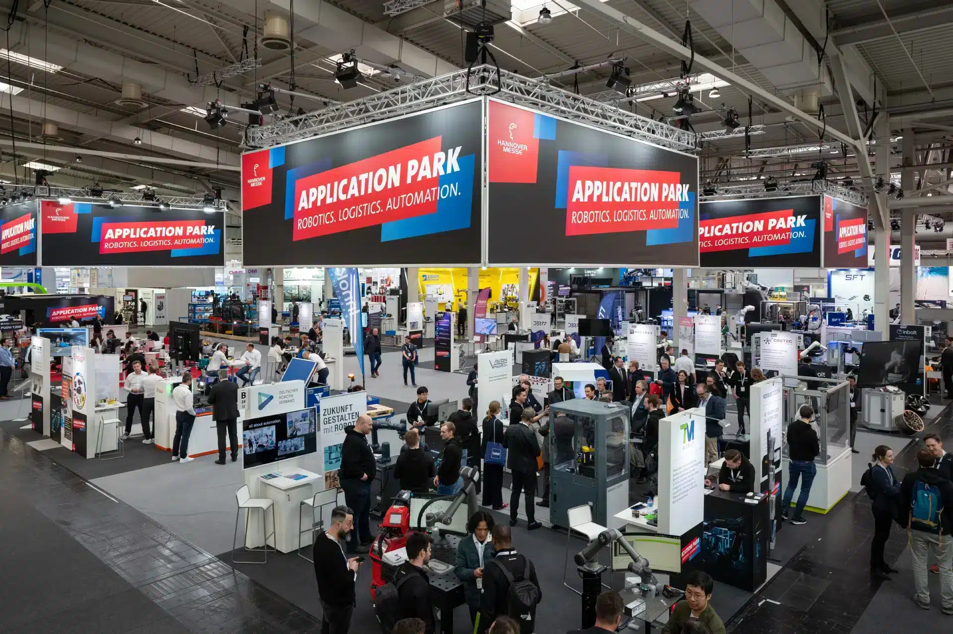 Auf der belebten Messefläche der Messe Hannover tummeln sich zahlreiche Stände, an denen Besucher interagieren und die Exponate erkunden. Über ihnen prangt ein großes Schild mit der Aufschrift „Application Park: Robotik, Logistik, Automatisierung“ – ein Hinweis auf den Technologieschwerpunkt der Veranstaltung.