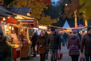 Menschen in Winterkleidung schlendern über den festlichen Freiluftmarkt der Messe Heilbronn. Lichtergeschmückte Holzstände bieten verschiedene Waren an. Bäume mit gelben Blättern und Lichterketten sorgen mit Einbruch der Dunkelheit für eine warme, einladende Atmosphäre.