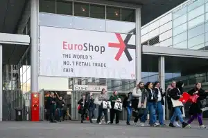Besucher schlendern vor einem modernen Kongresszentrum unter einem großen EuroShop-Banner hindurch, das die Messe als „weltweit führende Einzelhandelsmesse“ auszeichnet. Die glänzende Glas- und Metallkonstruktion ragt über den Besuchern empor, während Menschen mit roten und weißen Taschen in diesem Einkaufsparadies zusammenströmen.