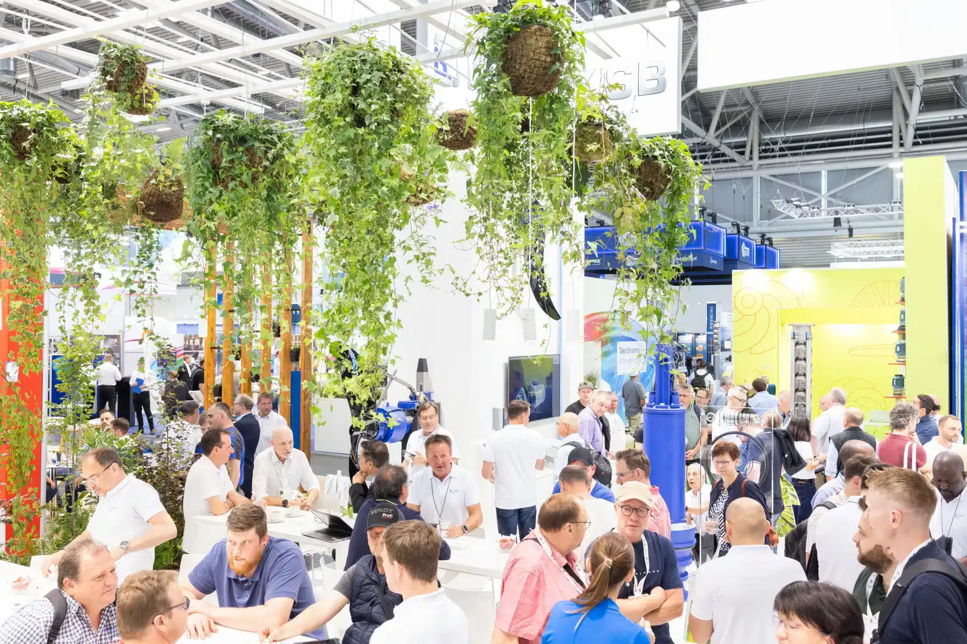 Eine geschäftige Indoor-Ausstellung erinnert an die IFAT, mit zahlreichen Menschen, die an Tischen interagieren. Üppige grüne Pflanzen hängen von der Decke und verleihen der hellen, geschäftigen Umgebung ein natürliches Element. Im Hintergrund sind verschiedene Geräte und farbenfrohe Displays zu sehen.