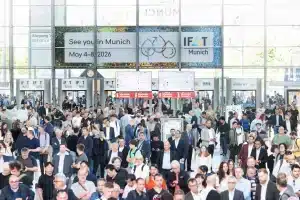In der Messehalle der IFAT München versammelt sich eine geschäftige Menschenmenge. Schilder mit der Aufschrift „Wir sehen uns in München, 4. bis 8. Mai 2026“ zieren das lebhafte Bild. Zahlreiche Menschen gehen geschäftig umher und interagieren angeregt.