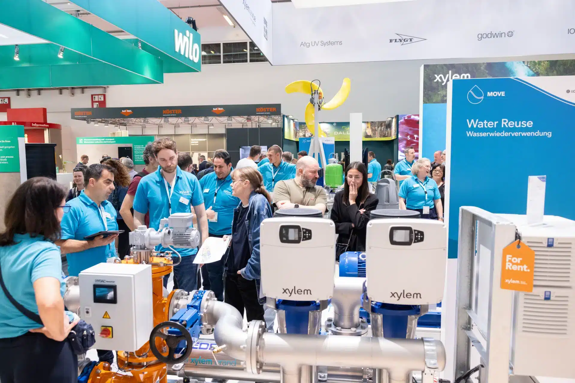 Eine Gruppe von Menschen in blauen Hemden steht auf der IFAT um Industrieanlagen herum. Am Stand sind Schilder für Wasserwiederverwendungstechnologie zu sehen, die das Interesse der Besucher wecken, die die Maschinen gespannt begutachten.