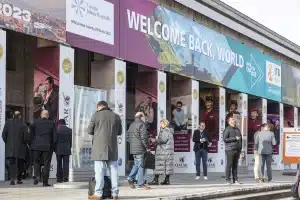 Menschen stehen vor einem Gebäude, auf dem ein großes Banner mit der Aufschrift „WILLKOMMEN ZURÜCK, WELT“ prangt – als Teil der ITB Berlin. Verschiedene Anzeigen und Logos deuten darauf hin, dass es sich um eine Ausstellung handelt, auf der Besucher sich unterhalten, umherschlendern und die geschäftige Atmosphäre genießen.