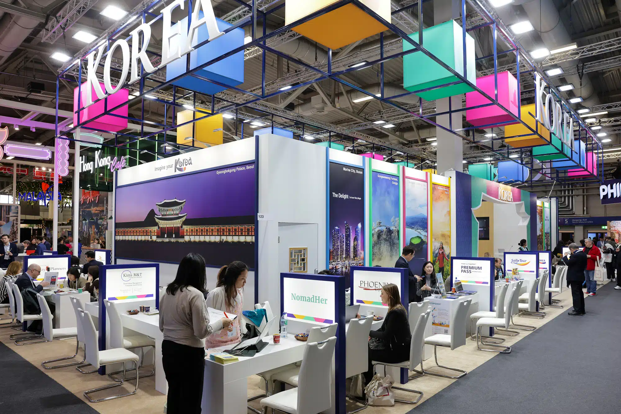 Ein Messestand auf der ITB Berlin präsentiert Korea-Themen. Besucher sitzen an Tischen mit Computern und diskutieren. Der Stand ist mit bunten quadratischen Lichtern und großen Schildern geschmückt, die für verschiedene koreanische Marken und Attraktionen werben und Besucher aus aller Welt begeistern.