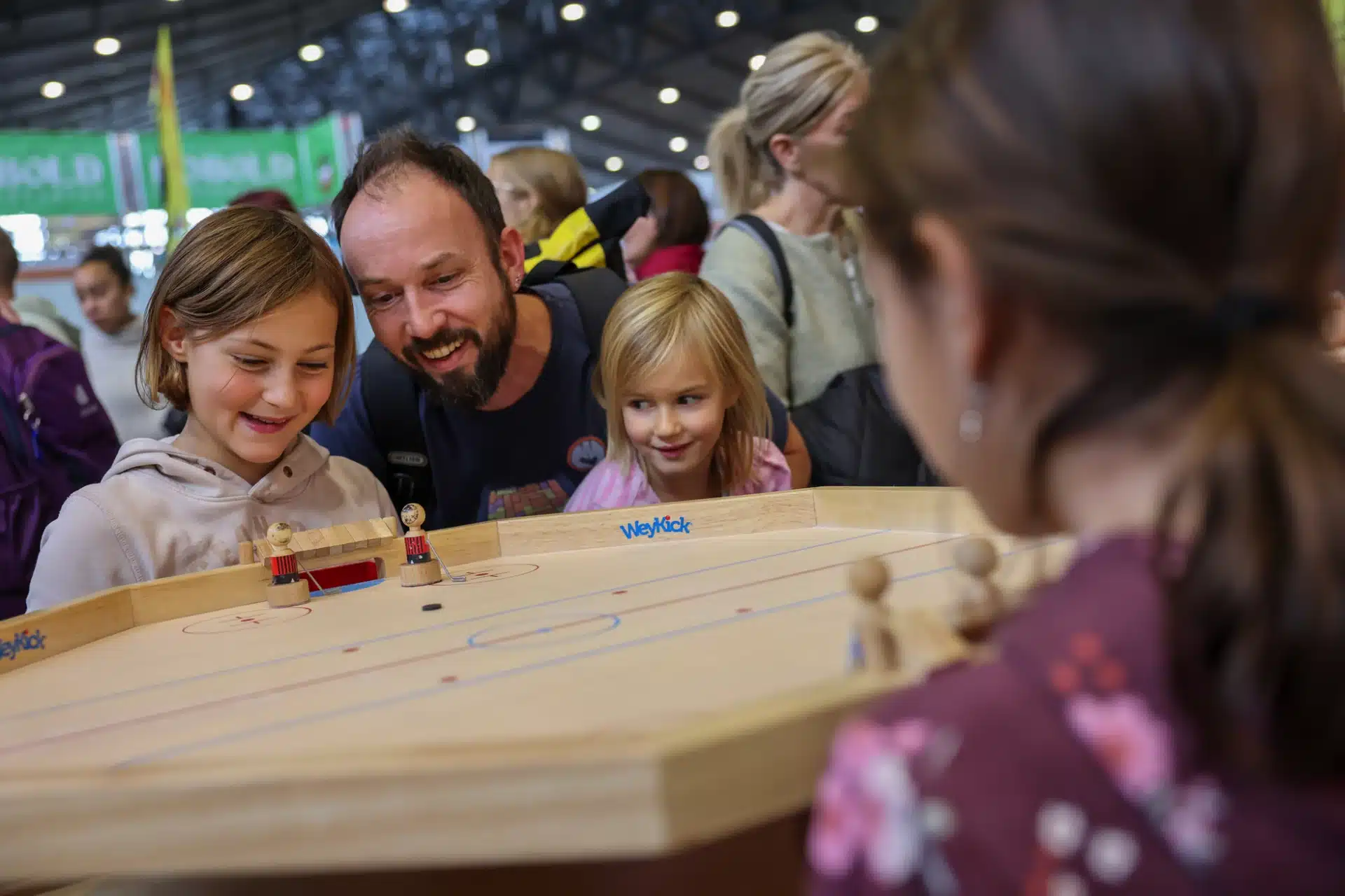 Eine Gruppe von Menschen schaut sich beim lebhaften Stuttgarter MesseHerbst ein Brettspiel an. Zwei Kinder und ein Mann lächeln und beobachten konzentriert den Minihockey-Aufbau. Die Atmosphäre ist fröhlich und anregend, und im Hintergrund amüsieren sich die anderen Besucher.