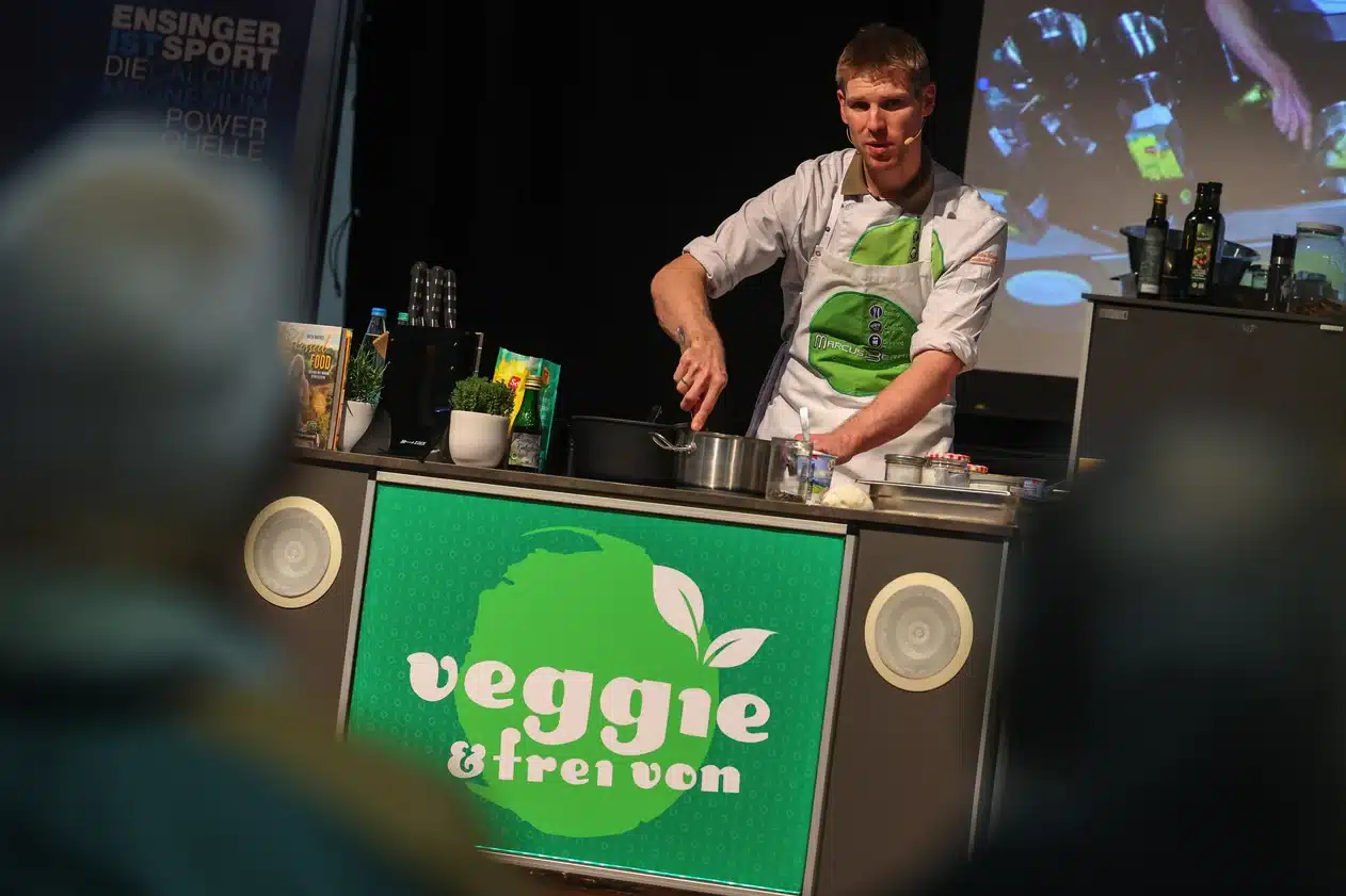 Beim Stuttgarter MesseHerbst kocht ein Mann in weißem Hemd und grüner Schürze an einer Vorführtheke mit der Aufschrift „veggie & frei von“. Hinter Töpfen und Kochgeschirr hängt ein Bildschirm mit Bildern köstlicher Gerichte.