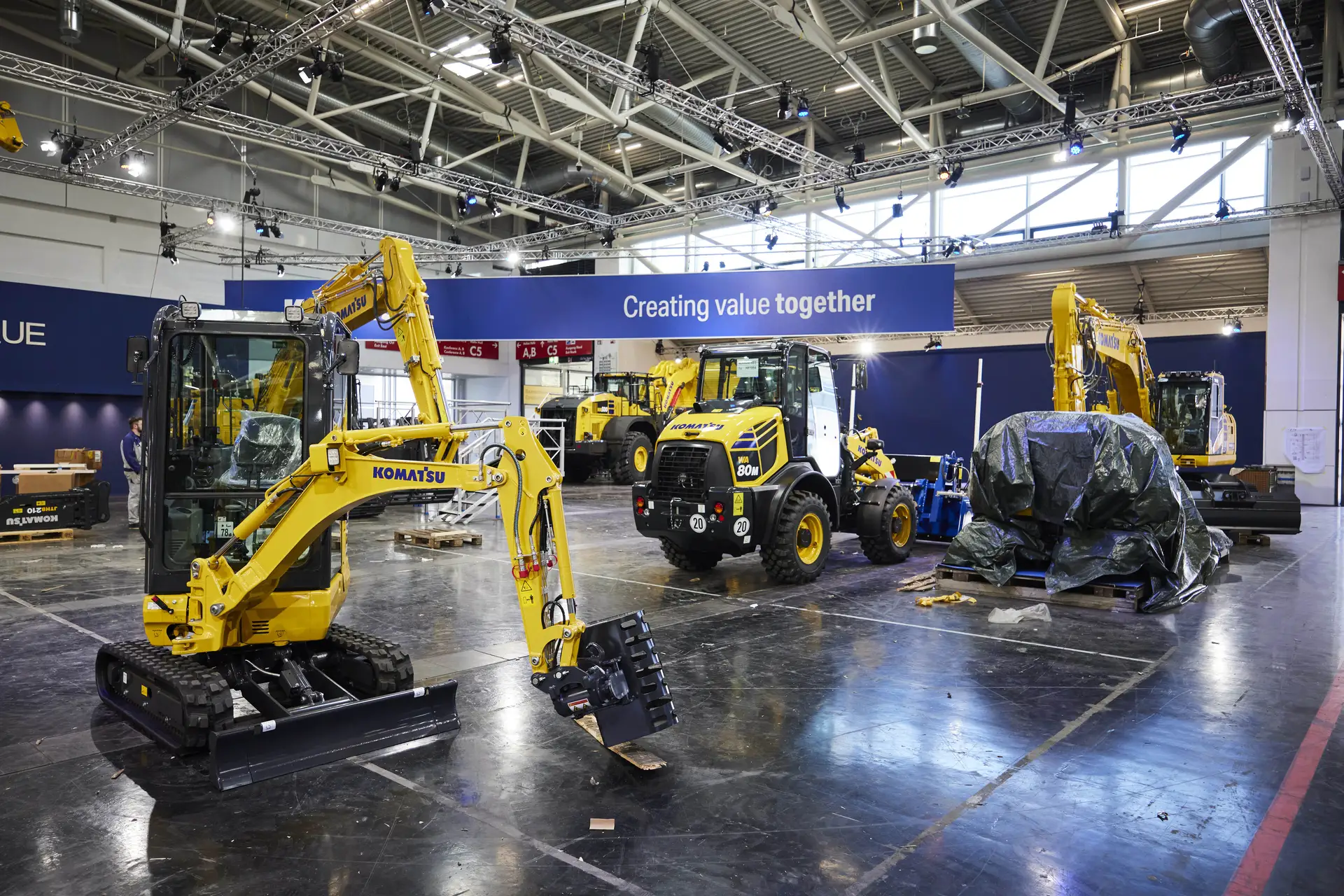 In der bauma-Halle werden Baumaschinen präsentiert. Mehrere gelbe Bagger und Maschinen sind unter hellem Licht aufgestellt, unter einem großen Banner mit der Aufschrift „Gemeinsam Werte schaffen“.