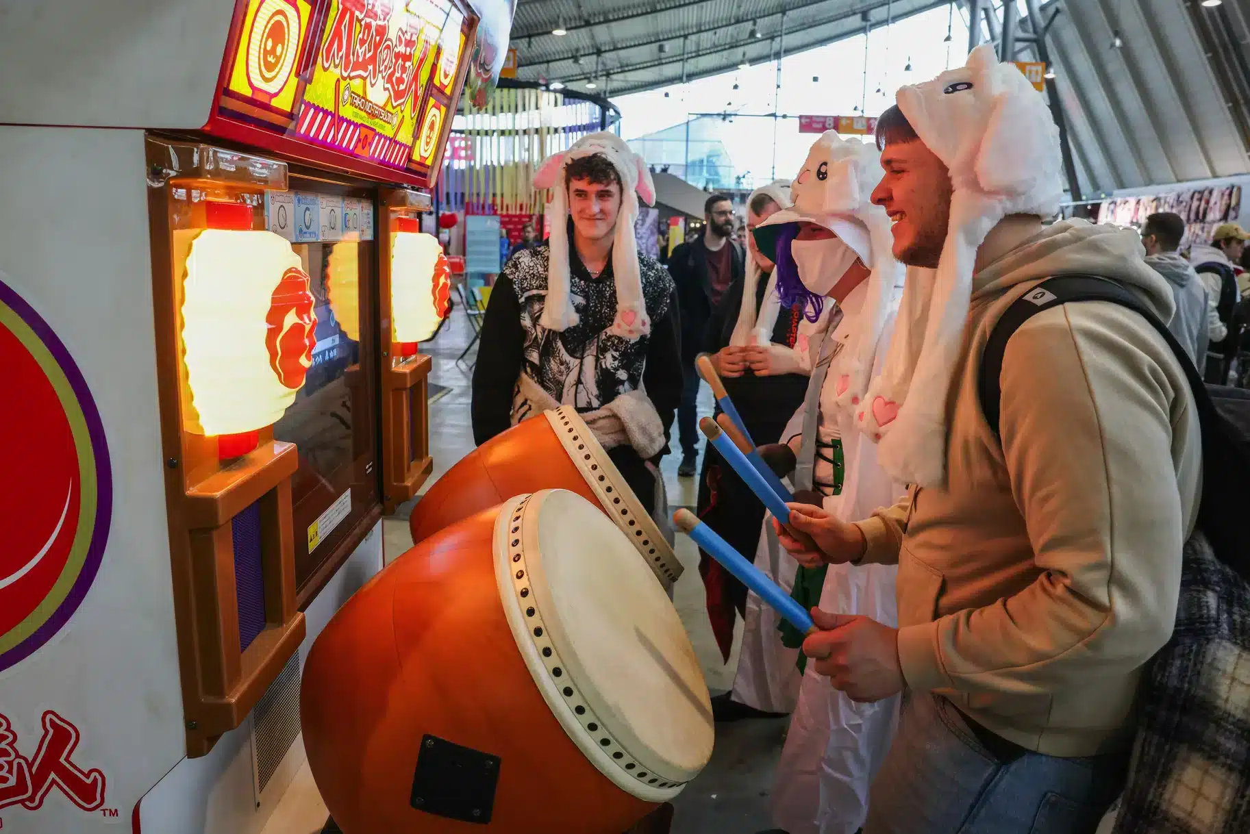 In einer Indoor-Veranstaltung spielen Menschen mit Tierhüten fröhlich ein Taiko-Trommel-Arcade-Spiel. Ihr Lächeln erhellt die Szene und erinnert an die lebhafte Atmosphäre einer Comic-Con. Im Hintergrund sind weitere Spielautomaten und Besucher zu sehen.