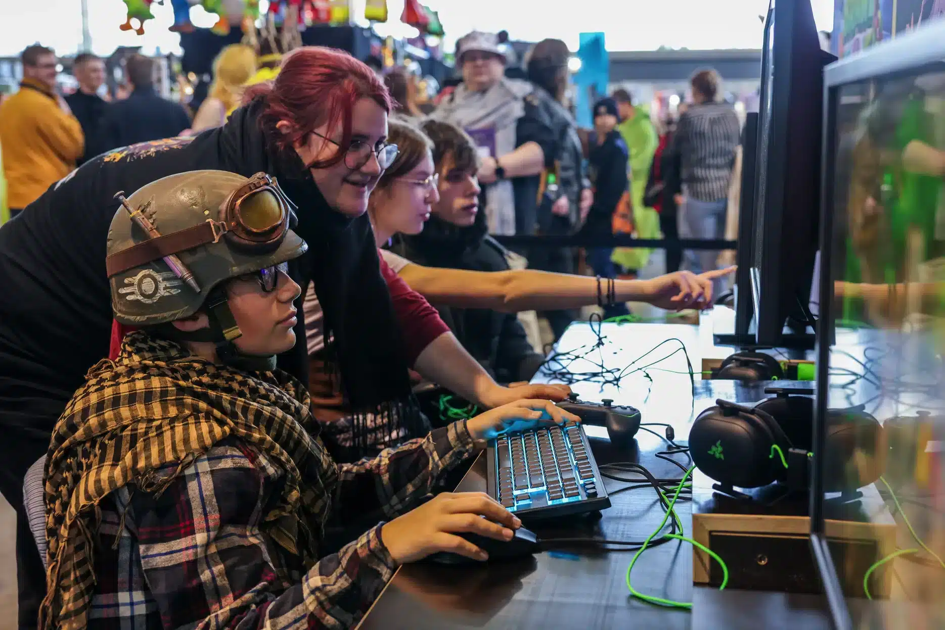 Auf der Comic Con wimmelt es im geschäftigen Messezentrum von Gamern an einem belebten Stand. Konzentrierte Spieler, darunter einer mit Helm und Schutzbrille, tippen auf Tastaturen. Währenddessen beugt sich ein rothaariger Helfer vor, um zu helfen, während farbenfrohe Waren den Hintergrund schmücken.