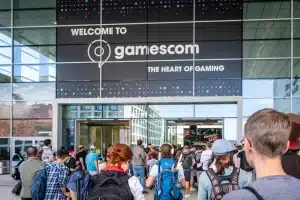Eine lebhafte Menschenmenge strömt in ein großes Gebäude und wird von dem Schild „Willkommen zur Gamescom – dem Herzen der Gaming-Szene“ begrüßt. Der Eingang mit seinen Glastüren und hohen Fenstern heißt die Besucher – viele mit Rucksäcken – herzlich willkommen, während sie in die pulsierende Welt der Gamescom eintauchen.