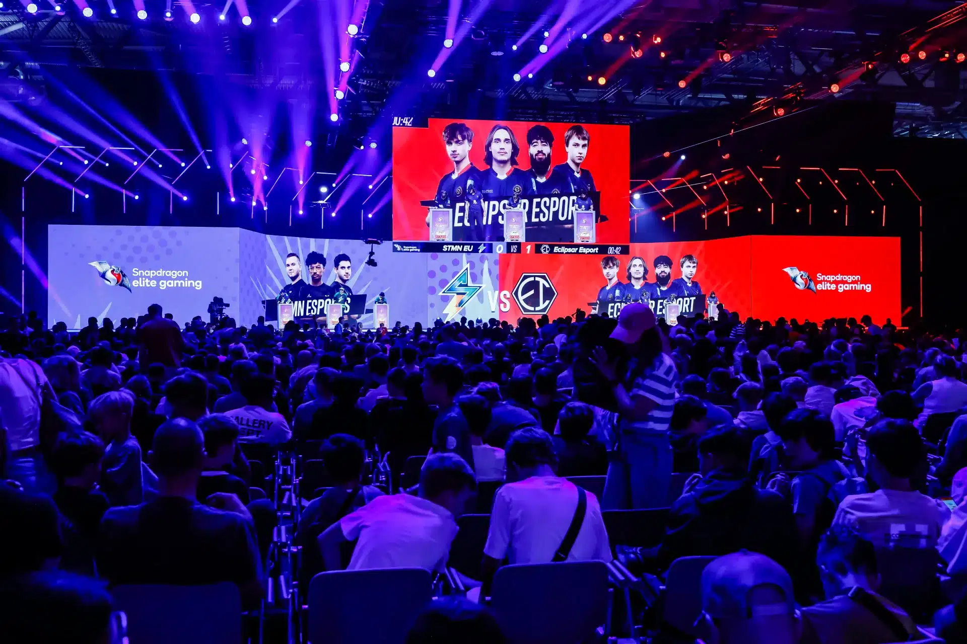 Eine große Menschenmenge verfolgt ein E-Sport-Event auf der Gamescome auf riesigen Bildschirmen in einer dunklen, lebendigen Arena. Teamgrafiken und Logos werden prominent auf den Bildschirmen angezeigt. Helle Bühnenlichter erhellen den Veranstaltungsort und sorgen für eine elektrisierende Atmosphäre.