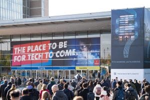Vor einem Kongresszentrum versammelt sich eine Menschenmenge. Große Banner mit der Aufschrift „The Place to Be for Tech to Come“ (Der Ort, an dem die Technologiebranche heute ihren Platz hat) erinnern an die Hannover Messe. Das Gebäude verfügt über mehrere Eingänge und eine digitale Anzeige für Cisco an der Gebäudeseite.