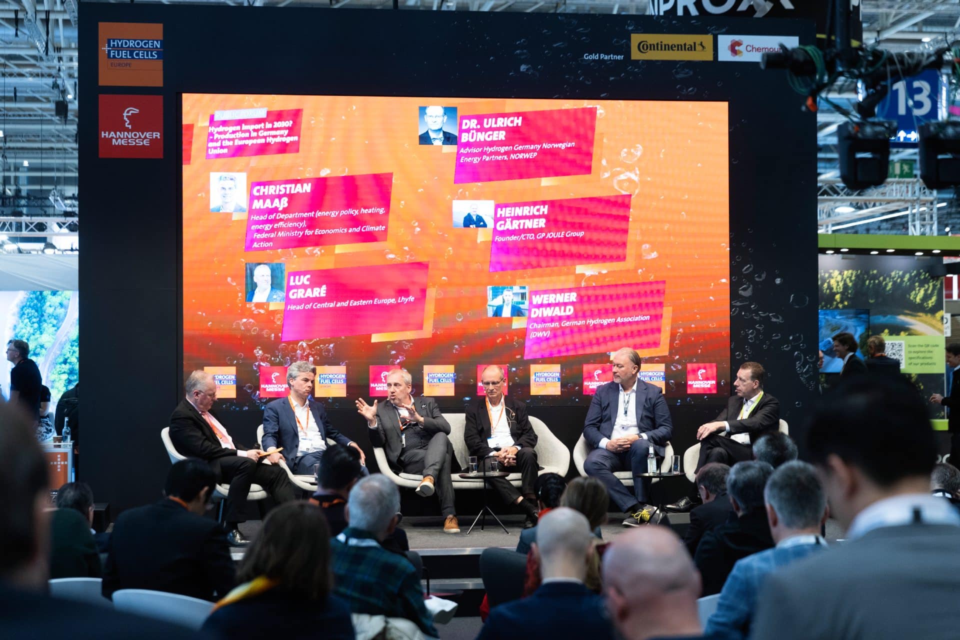 Ein Panel aus sechs Männern in Businesskleidung sitzt auf einer Bühne der Hannover Messe vor einer großen Leinwand mit Namen und Titeln. Im Vordergrund sitzt ein Publikum und verfolgt die Diskussion. Der Veranstaltungshintergrund zeigt Markenlogos und Energiethemen.