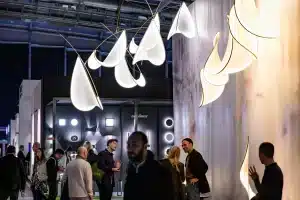 Menschen versammeln sich in einem Ausstellungsraum der Light + Building. Moderne, blattförmige Leuchten hängen von der Decke. Im Hintergrund sind dunkle Wände mit kreisförmigen Lichteffekten zu sehen. Einige unterhalten sich, während andere den Raum erkunden.
