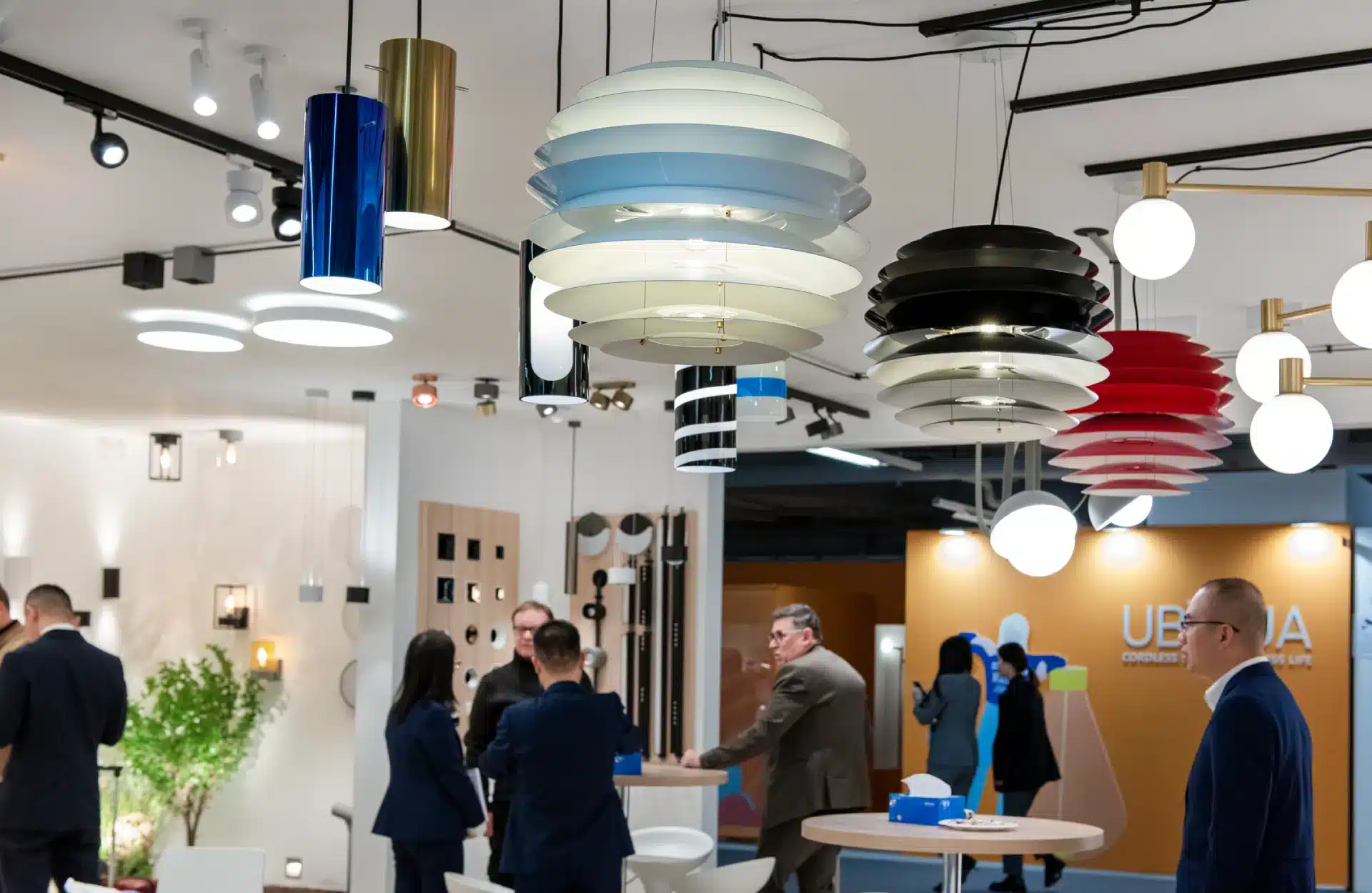 Ein moderner Showroom präsentiert verschiedene Pendelleuchten in unterschiedlichen Farben und Designs, die den Raum brillant erhellen. Vor dem Hintergrund dieser einladenden Lichtlandschaft unterhalten sich Menschen in Anzügen an einem Tisch, während ein Schild mit der Aufschrift „UBE“ den architektonischen Geist der Light + Building widerspiegelt.