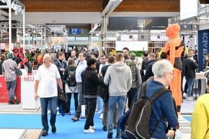 Die lebhafte Atmosphäre der Messe Kassel ist pulsierend, während eine bunt gemischte Gruppe durch die Ausstellungshalle strömt. Stände und Ausstellungen säumen beide Seiten, wobei eine leuchtend orange Schaufensterpuppe prominent auf der rechten Seite steht und alle Blicke auf sich zieht.