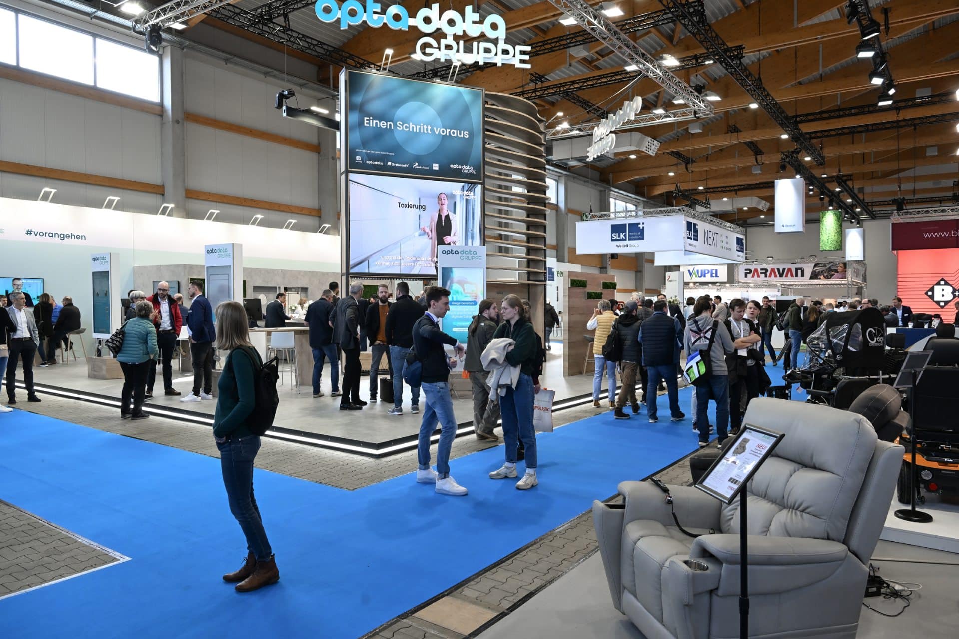 Auf der Messe Kassel herrscht reges Treiben und Geplauder. An den Ständen werden verschiedene technische Exponate unter hellem Licht präsentiert. Ein großes Banner trägt die Aufschrift „opta data gruppe“. Ein blauer Teppich führt die Besucher durch die Messe, wo bequeme Sitzgelegenheiten in einem Bereich angeordnet sind.