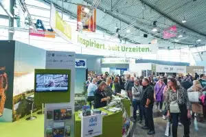 Eine geschäftige CMT-Messe mit Besuchern an verschiedenen Ständen. Das Schild über der Messe trägt die Aufschrift „Inspiriert. Seit Urzeiten“. Besucher unterhalten sich mit Ausstellern, und Broschüren und Informationen sind sichtbar. Die Atmosphäre ist lebendig und gut beleuchtet.