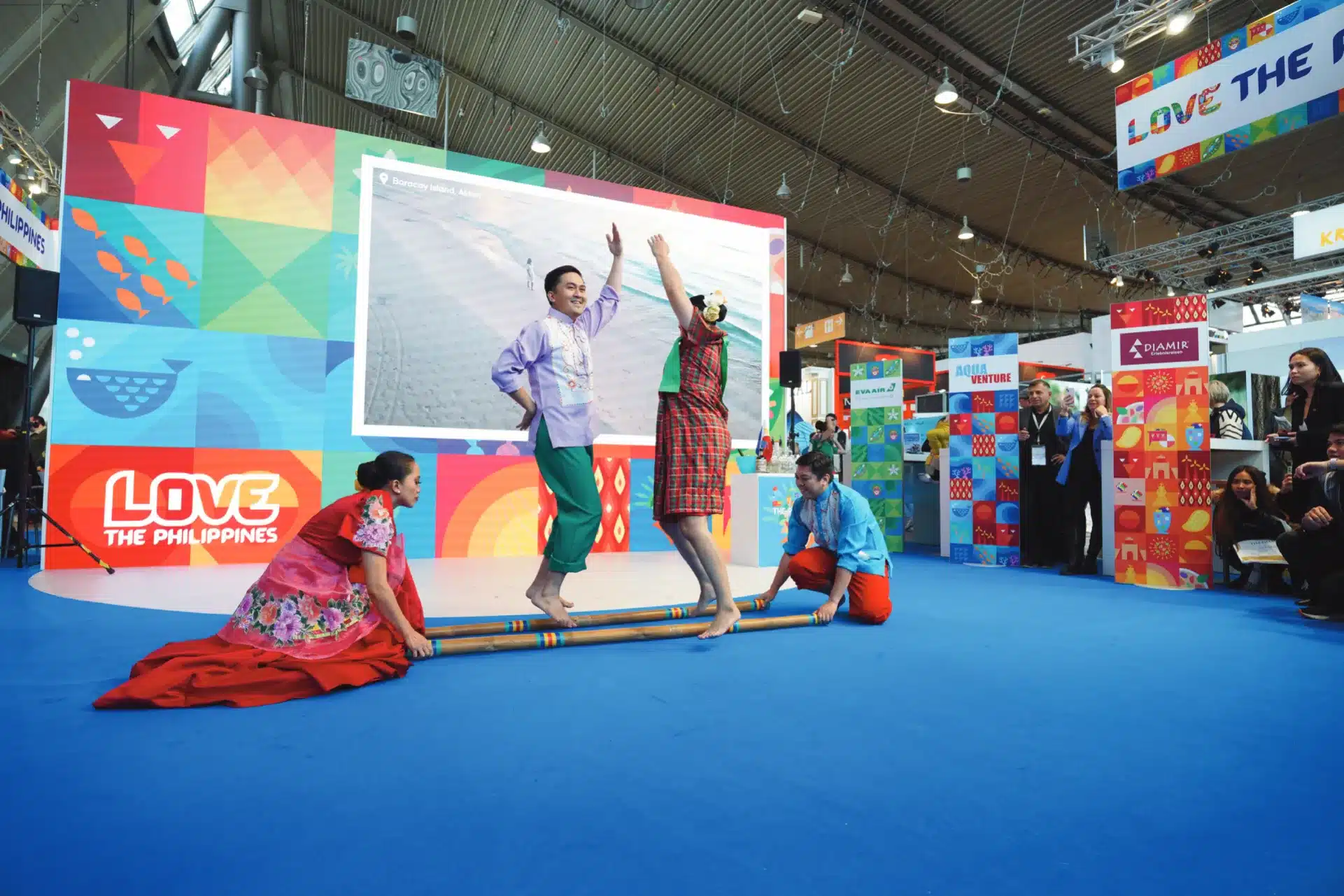 Künstler in farbenfroher traditioneller Kleidung tanzen mit Bambusstäben im Rahmen einer kulturellen Darbietung beim CMT-Festival. Auf einer Leinwand ist eine malerische Kulisse zu sehen, bunte Banner schmücken den Bereich. Zuschauer beobachten den Aufführungsort.
