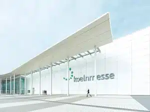 Die Außenansicht des Kölnmesse-Gebäudes besticht durch seine moderne weiße Fassade und die große Beschilderung. Drei Personen stehen links am Eingang, eine weitere Person geht im Vordergrund. Die Szene spielt vor dem Hintergrund eines strahlend blauen Himmels.