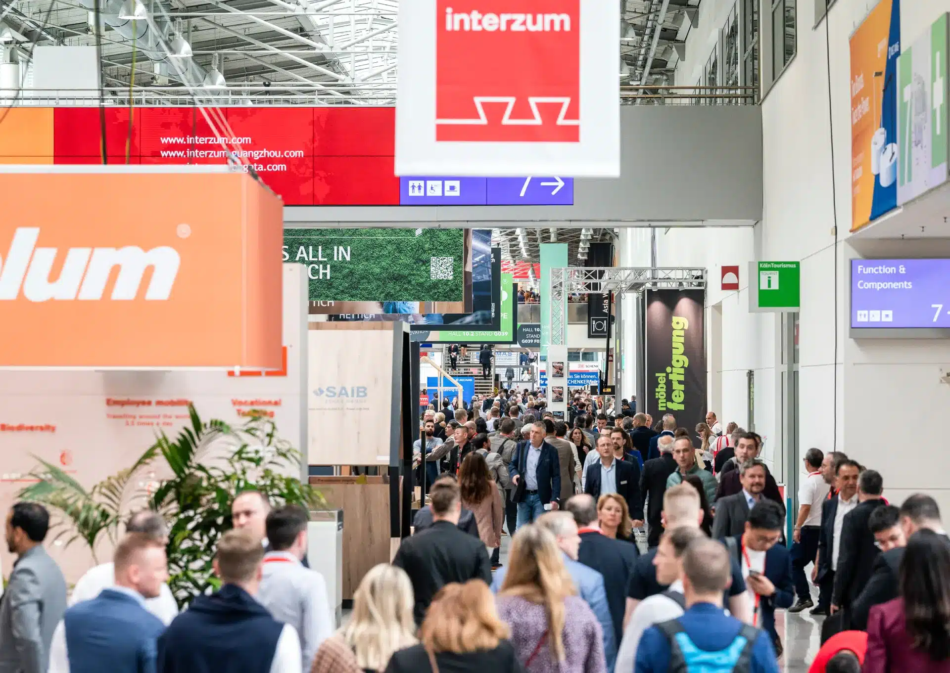 Auf der Kölnmesse herrscht reges Treiben mit Besuchern unterschiedlichster Herkunft. Farbenfrohe Stände und Beschilderungen, darunter ein markantes „Interzum“-Schild, prägen den Raum. Die Atmosphäre ist lebhaft, Networking und die Erkundung der Messe sind in vollem Gange.