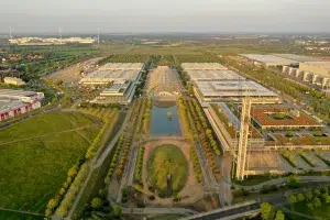 Aus der Luft betrachtet präsentiert sich das weitläufige Messegelände Leipzig mit einem zentralen Gebäude mit Glaskuppel, umgeben von symmetrisch angelegten Grünflächen und Wegen. Reflektierende Wasserspiele ergänzen das Design, während Windräder anmutig in der Ferne in die Landschaft hineinragen.