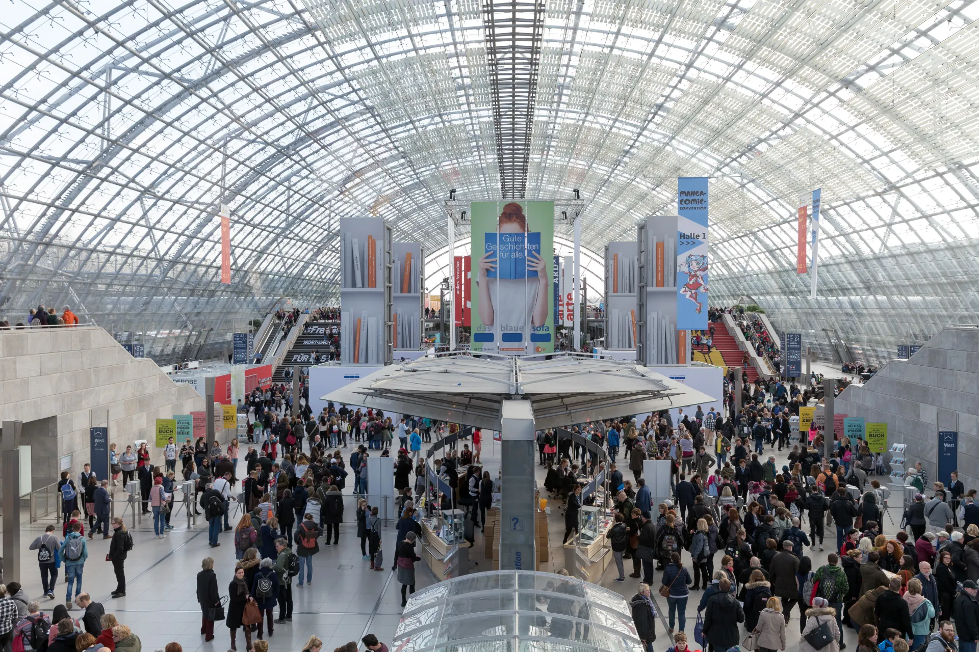 Ein großes, geschäftiges Messegelände mit hohem Glasdach spiegelt die Energie der Messe Leipzig wider. Eine Menschenmenge bewegt sich zwischen Ausstellungen und Ständen, während leuchtende Banner und Schilder darüber wehen. Im Hintergrund fahren Rolltreppen durch das pulsierende Treiben nach oben.