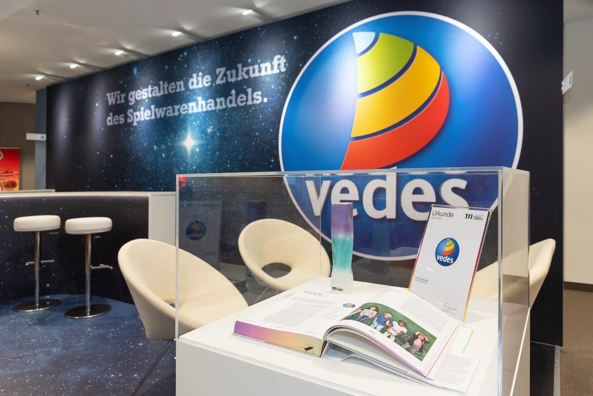 Messebau Nürnberg – Vedes Präsentationsbereich mit Urkunde und Magazin im Vordergrund sowie modernem Messestand auf der Spielwarenmesse 2025.