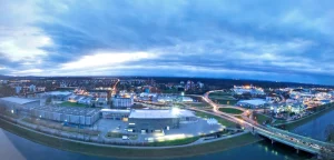 Panoramablick auf eine Stadtlandschaft in der Abenddämmerung mit beleuchteten Straßen und Kreuzungen unter einem bewölkten Himmel. Gebäude und Infrastruktur sind sichtbar. Im Vordergrund verläuft ein Fluss, über den eine Brücke in der Nähe der Messe Offenburg führt.