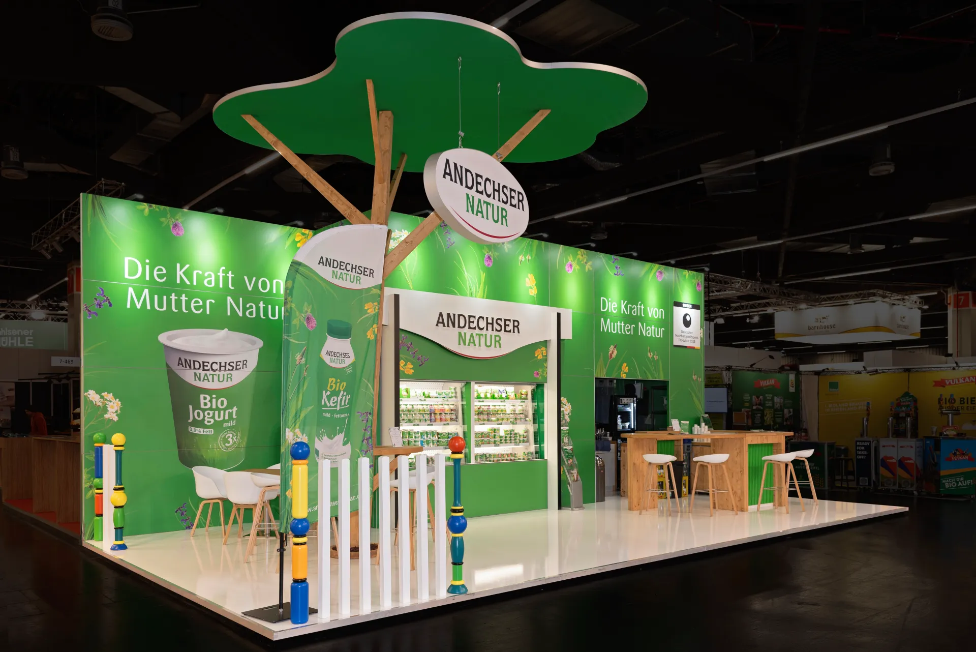 Andechser Natur Messestand mit leuchtendem Baumdach, Holztheke und Produktpräsentation auf der BioFach Messe 2025 in Nürnberg.