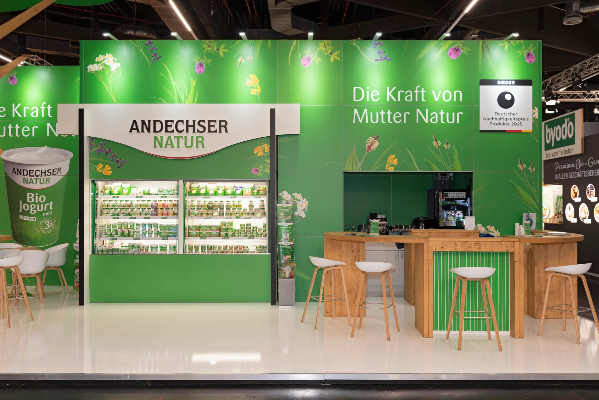 Frontansicht des Andechser Natur Messestandes mit Bio-Joghurt-Kühlregal und Holzbar auf der BioFach Messe 2025 in Nürnberg.