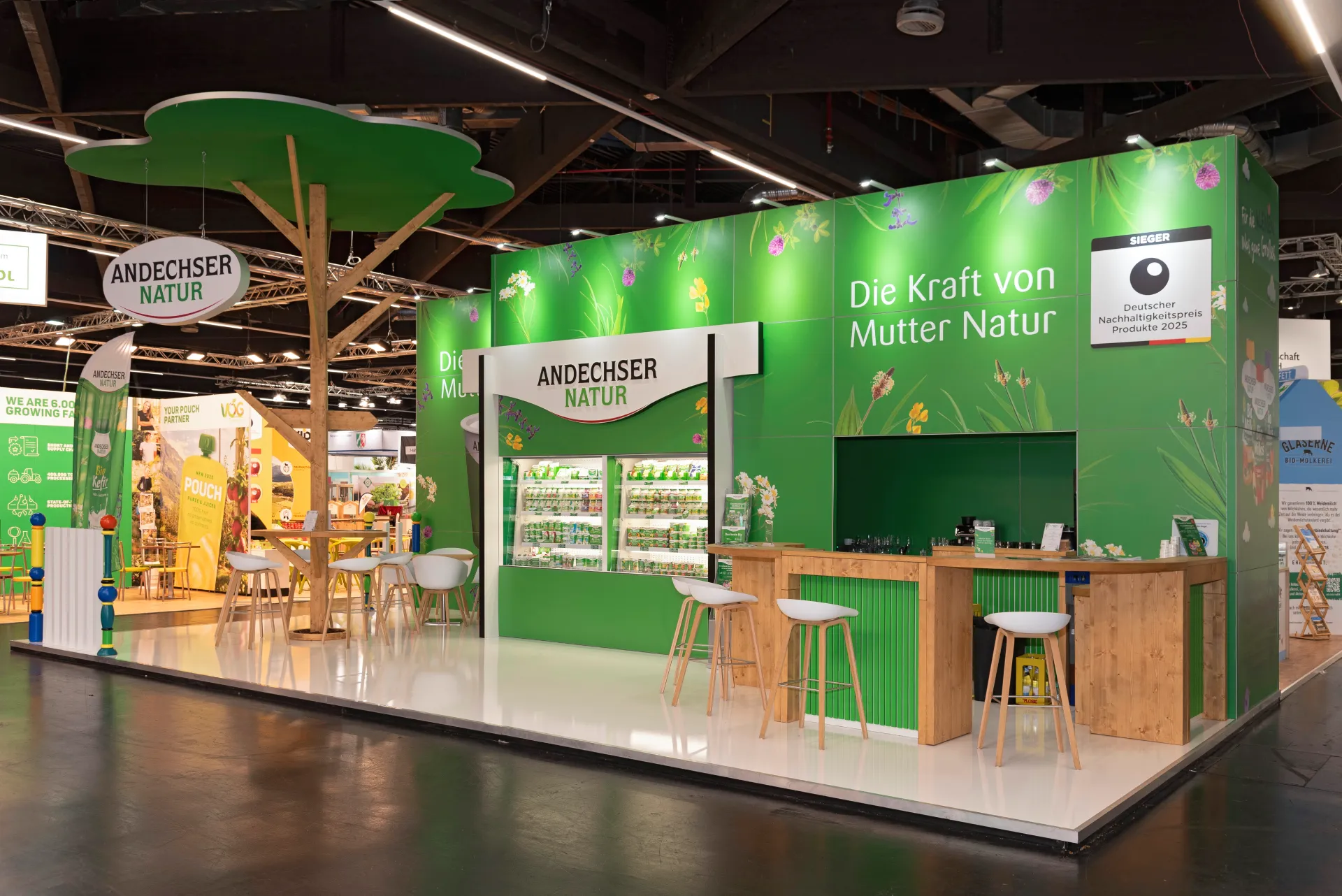 Andechser Natur Messestand mit zentralem Baum-Design, Holztheke und nachhaltiger Präsentation auf der BioFach Messe 2025 in Nürnberg.