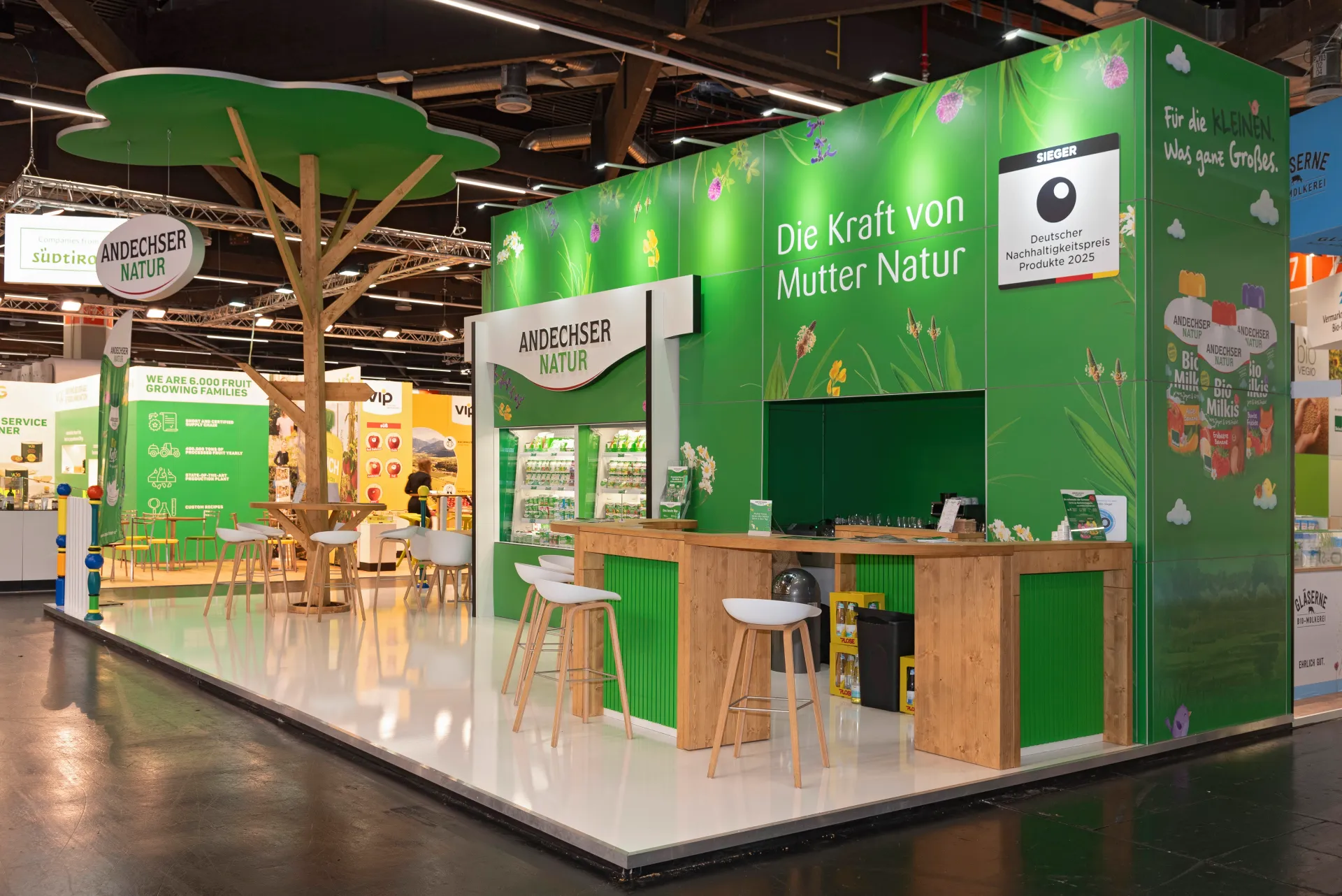 Grüner Messestand von Andechser Natur mit Holztheke, Sitzgelegenheiten und großem Baum-Element auf der BioFach Messe 2025 in Nürnberg.