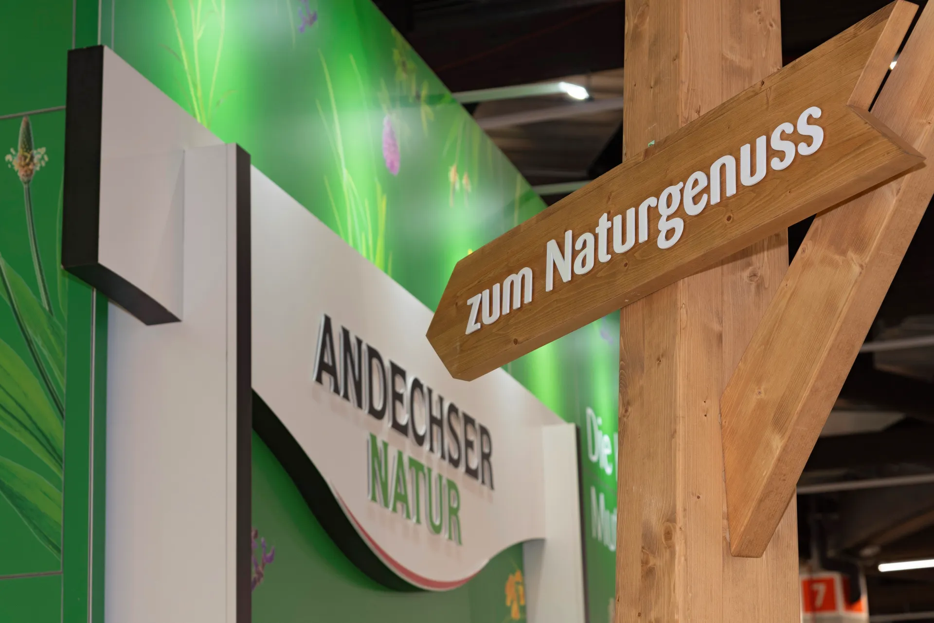 Holzwegweiser mit Aufschrift „zum Naturgenuss“ vor dem Andechser Natur Messestand auf der BioFach Messe 2025 in Nürnberg.